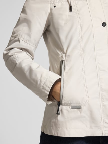 Kobiece model nosi Weather Protection Jack mit Reißverschlusstaschen od Gil Bret w kolorze jasnobeżowym, krój regular fit, kaptur z tunelem, stójka, zamek błyskawiczny, listwa guzikowa, kieszenie zapinane na zamek błyskawiczny, długość tyłu w rozmiarze 36: 66 cm, szerokość ramion w rozmiarze 36: 40 cm, szerokość w rozmiarze 36: 48 cm, długość rękawów w rozmiarze 36: 63 cm, zdjęcie: zbliżenie górnej części ciała z przodu.