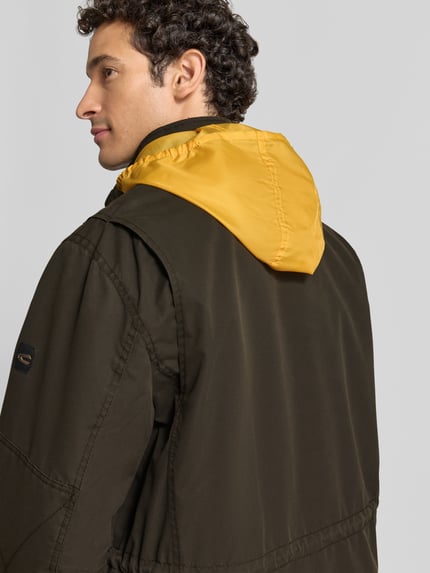 Männermodel trägt Jacke mit Stehkragen und Pattentaschen von camel active in Olivgrün mit gelber Kapuze, Ganzkörperansicht Rückansicht. Rückenlänge 79 cm, Schulterbreite 44 cm, Brustweite 65 cm, Ärmellänge 71 cm, Regular Fit, Knopfleiste und Ärmelriegel, glatte Textur.