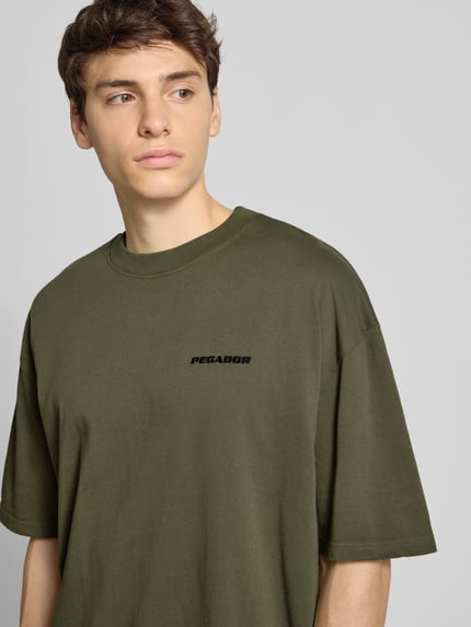 Männermodel draagt Oversized T-Shirt mit Label-Stitching van Pegador, katoen, olijfgroen, geribde ronde hals, Nahaufnahme bovenlichaam.