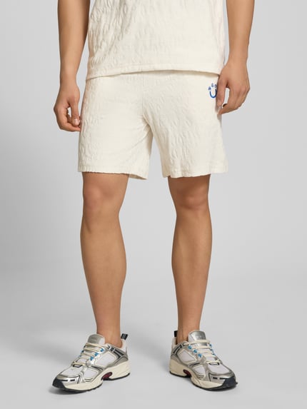 Männermodel trägt Regular Fit Shorts aus Frottee von HUGO in Creme mit Frottee-Optik, Logo-Stitching, Tunnelzug, seitlichen Eingrifftaschen, aufgesetzter Tasche hinten, Ganzkörperansicht von vorne.