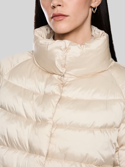 Frauenmodel trägt Steppjacke mit Stehkragen und Label-Detail von Marc Cain in Beige, glänzende Oberfläche, Nahaufnahme Oberkörper. Damen Steppjacke, Regular Fit, eingearbeitete Taschen, Länge 56 cm, Schulterbreite 41 cm, Brustweite 55 cm, Ärmellänge 73 cm.