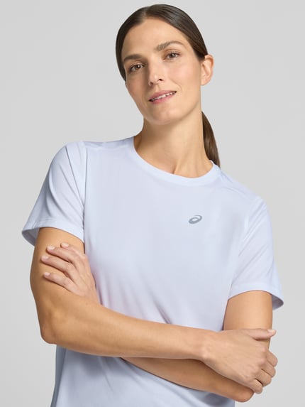 Frauenmodel trägt T-Shirt mit Rundhalsausschnitt und Logo-Print von ASICS in Weiß, glatte Struktur, Nahaufnahme Oberkörper, gerader Schnitt, leicht abgerundeter Saum, Rückenlänge Größe S 60 cm.