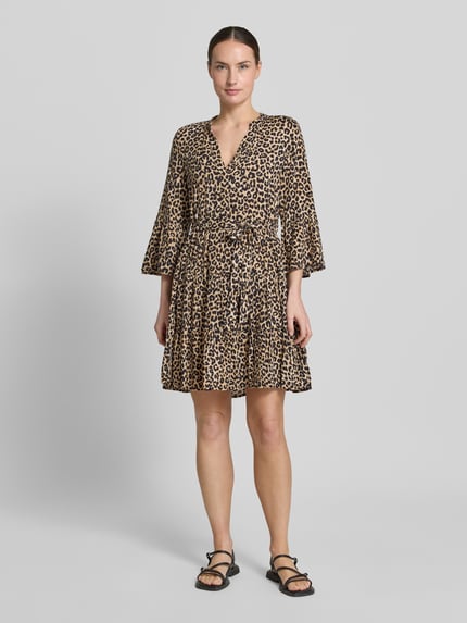 Frauenmodel trägt tailliertes Tunikakleid aus reiner Viskose Modell 'MELBA' von Vero Moda mit Allover-Muster, V-Ausschnitt, Bindegürtel, Mini-Länge, Volantsaum, Trompetenärmeln, in Beige mit Brauntönen, leichte Stoffstruktur, Ganzkörperansicht von vorne.