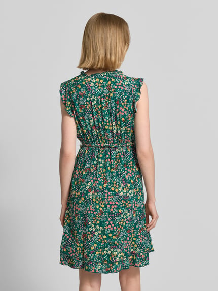Vrouwenmodel draagt mini-jurk uit zuiver viscose met bloemen all-over print van Apricot, getailleerd, met ruches, mouwloos, afneembare tailleband, V-hals, knoopsluiting, ruglengte maat S 85 cm, borstwijdte 30 cm, mouwlengte 6 cm, geheel groene, kleurrijke bloemenprint, gladde stof, getextureerde ruches, geheelopname rugaanzicht.