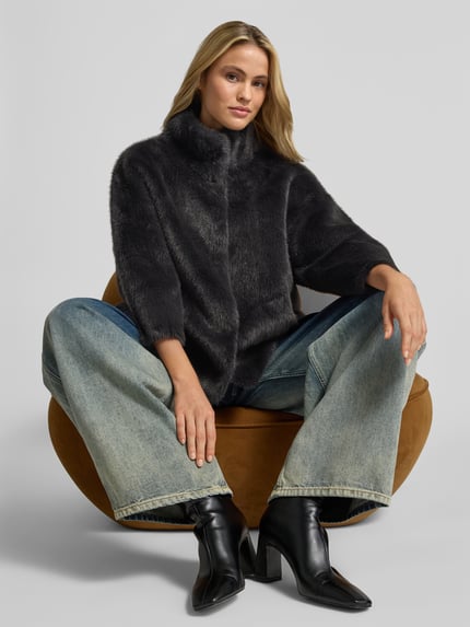Modelka kobieca ma na sobie Wendejacke aus Kunstfell od Juvia, prosty krój, sztuczne futro, funkcja dwustronna, stójka, wpuszczane kieszenie, długość tyłu w rozmiarze S 63 cm, szerokość w rozmiarze S 54 cm, długość rękawów w rozmiarze S 47 cm, ciemne odcienie, gładka tekstura, całkowity widok z przodu.