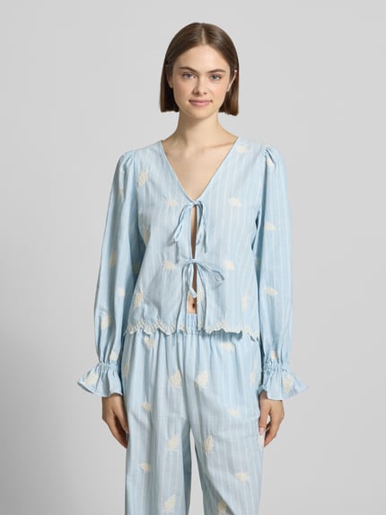 Vrouwenmodel draagt blouse met vetersluiting Modell 'Ninon' van OH APRIL in lichtblauw met witte print van puur katoen, straight fit, V-hals, labelstitching, mouwen met ruches, ruglengte maat S 54 cm, mouwlengte maat S 61 cm. Ganzekörperaanzicht van voren.