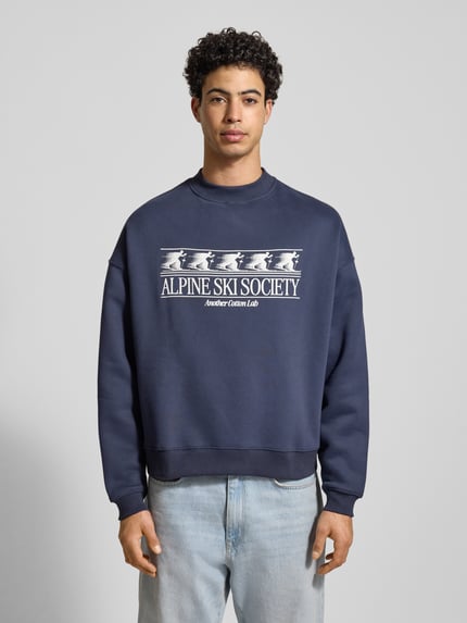 Męski model nosi Sweatshirt mit Label-Print od ANOTHER COTTON LAB, mieszanka bawełny, luźny krój, prążkowane wykończenia, okrągły dekolt, nadruk z logo, ciemnoniebieski kolor, widok całej sylwetki z przodu.