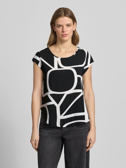 Frauenmodel trägt Relaxed Fit Blusenshirt in Crêpe-Verarbeitung Modell 'VIC' von Only mit Optik:Allover-Muster, U-Boot-Ausschnitt, kurzem Reißverschluss, Kappärmel, abgerundetem Rumpfabschluss, Materialverarbeitung: Crêpe in Schwarz und Weiß mit geometrischen Formen, Ganzkörperansicht von vorne.