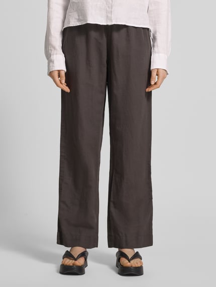 Frauenmodel trägt Stoffhose mit Leinen-Anteil und Eingrifftaschen von Neo Noir, dunkler Baumwoll-Leinen-Mix, ausgestellter Schnitt, Ganzkörperansicht von vorne. Damen Stoffhose, Modell 'Sonar', elastischer Bund, Bundweite 36 cm, Innenbeinlänge 79 cm.