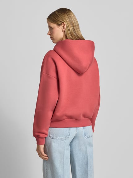 Modelka nosi Relaxed Fit Sweatjacke mit Kapuze und Logo-Print od REVIEW, mieszanka bawełny, krój relaxed fit, kaptur, nadruk z logo, zamek błyskawiczny, kieszeń kangurka, kolor różowy, wyrazista tekstura, gładka powierzchnia, widoczne przeszycia, widok całej sylwetki z tyłu