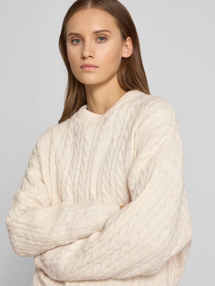Frauenmodel trägt Oversized Fit Strickpullover mit Zopfmuster von REVIEW in Creme, elastischer Material-Mix, Nahaufnahme Oberkörper.