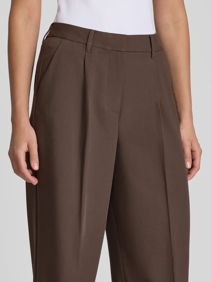 Kobieta nosi brązowe spodnie Wide Fit Hose mit Viskose-Anteil Modell 'Aya Ellis' od JJXX, z zakładkami i przeszyciami, materiał elastyczny, widoczne kieszenie, szerokość w rozmiarze S/32: 37 cm, długość nogawki wewnętrznej: 77 cm, zamek i haftka, zbliżenie na górną część spodni, zdjęcie przodu.