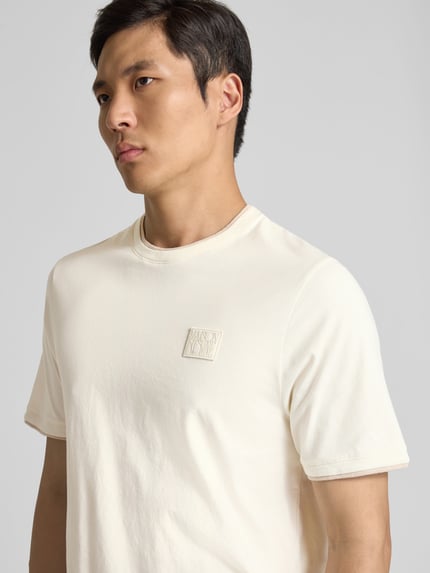 Mężczyznamodel nosi Slim Fit T-Shirt aus Baumwolle von MAISON KITSUNÉ, męski t-shirt, krój slim fit, bawełna, okrągły dekolt, detal z logo, wykończenia w kontrastowym kolorze, długość tyłu w rozmiarze M: 69 cm. Zbliżenie na górną część ciała z przodu.