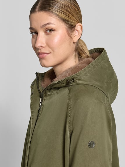 Kobieta nosi Parka mit Kapuze und Pattentaschen Modell 'EMMA' od Blonde No. 8, mieszanka bawełny z dodatkiem streczu, prosty krój, kaptur z tunelem i stoperami, zamek błyskawiczny i rzep, kieszenie z patką, patki przy mankietach, długość tyłu w rozmiarze 36: 105 cm, szerokość ramion w rozmiarze 36: 36 cm, szerokość w rozmiarze 36: 60 cm, długość rękawów w rozmiarze 36: 75 cm, oliwkowa, faktura matowa, zbliżenie na górną część ciała z przodu.