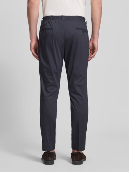 Männermodel trägt Regular Fit Stoffhose mit Logo-Detail Modell 'Lester' von JOOP! Collection aus dunkelblauem Baumwoll-Mix mit Bundfalten, Gesäßtaschen und Knopf-Reißverschluss, Ganzkörperansicht Rückansicht.