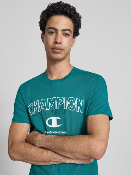 Männermodel trägt T-Shirt mit Label-Print und Rundhalsausschnitt von CHAMPION in Grün, reine Baumwolle, Regular Fit, Nahaufnahme Oberkörper.