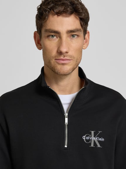 Männermodel trägt Regular Fit Sweatshirt mit Logo-Stitching Modell 'Terry' von Calvin Klein Jeans, Nahaufnahme Oberkörper, schwarze Baumwollmischung, dezenter Logo-Print, Troyer-Kragen mit Zipper, elastische Abschlüsse.