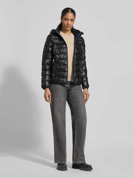 Frauenmodel trägt schwarze Jacke mit Kapuze von NAUTGÅRD, lockerer Schnitt, Tunnelzug, verdeckter Reißverschluss, Ganzkörperansicht von vorne.