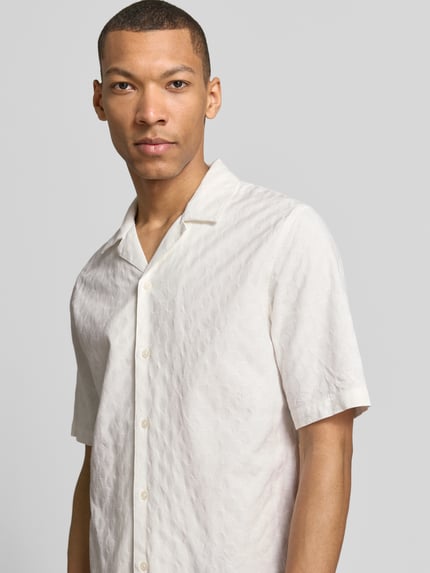 Męski model nosi koszulę casualową Relaxed Fit Freizeithemd mit Strukturmuster model 'OSCAR' od Samsøe Samsøe z bawełny, fakturowany wzór, kołnierz z połami, listwa guzikowa, kolor jasny kremowy, długość tyłu w rozmiarze M: 73 cm, szerokość ramion w rozmiarze M: 43 cm, długość rękawów w rozmiarze M: 30 cm, nahaufnahme górnej części tułowia.