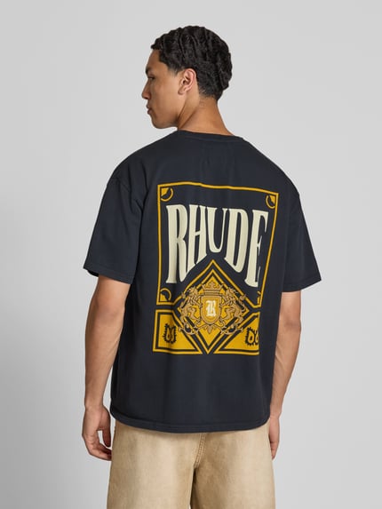 Männermodel trägt T-Shirt mit Label-Print von RHUDE, schwarze Farbe, Reine Baumwolle, Gerader-Schnitt, Kurzärmelig, Ganzkörperansicht Rückansicht.