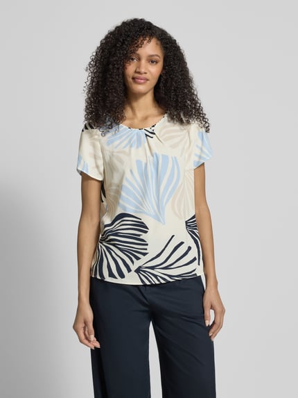 Modelka prezentuje Loose Fit Blusenshirt mit Allover-Muster von s.Oliver BLACK LABEL, krój loose fit, okrągły dekolt, wzór na całej powierzchni, kontrafałda, lekko zaokrąglone wykończenie, szyfon, jasne kolory z jasnoniebieskimi i czarnymi liśćmi, krótkie rękawy, efekt lekki, wykończenie materiału: szyfon. Ganzkörperansicht von vorne.