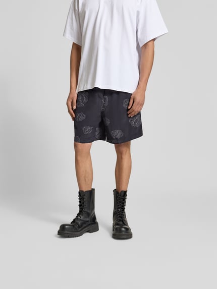 Männermodel trägt Shorts aus reiner Viskose von Officine Générale in Schwarz mit Muster, elastischem Bund und Tunnelzug, Ganzkörperansicht von vorne.