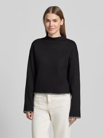 Frauenmodel trägt Pullover mit ausgestelltem Ärmelabschluss von JAKE*S WOMAN COLLECTION, unifarbener matt-schwarzer Strick, Cropped-Länge, hochgeschlossen mit Mock Neck, überschnittene lange Ärmel, abgerundeter Rumpfabschluss, Ganzkörperansicht von vorne.