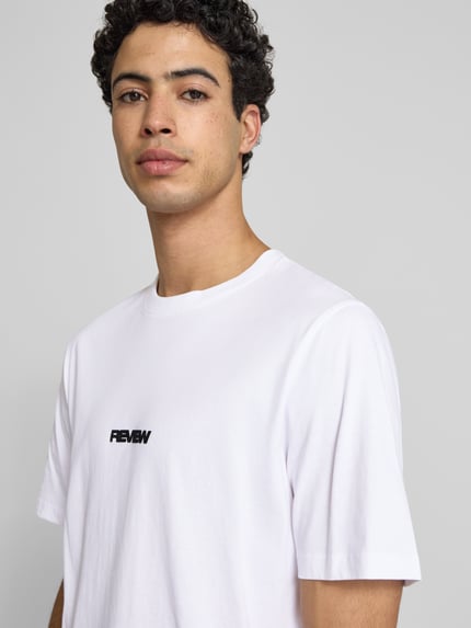 Männermodel trägt T-Shirt mit Logo und Rundhalsausschnitt von REVIEW in Weiß aus Reine Baumwolle, Relaxed-Fit, Nahaufnahme Oberkörper.