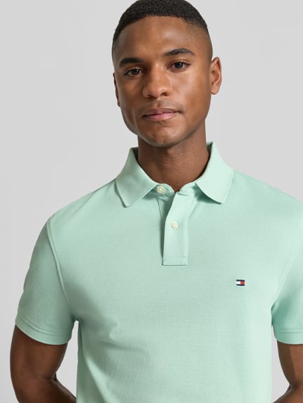 Mężczyznę model nosi Regular Fit Poloshirt aus Baumwoll-Mix von Tommy Hilfiger, efekt: jednolity kolor, krój regular fit, wyhaftowane logo, kołnierzyk polo, zapięcie na 2 guziki, krótki rękaw, wykończenie: z rozcięciem, wykończenie materiału: pika, mieszanka bawełny. Nahaufnahme Oberkörper.