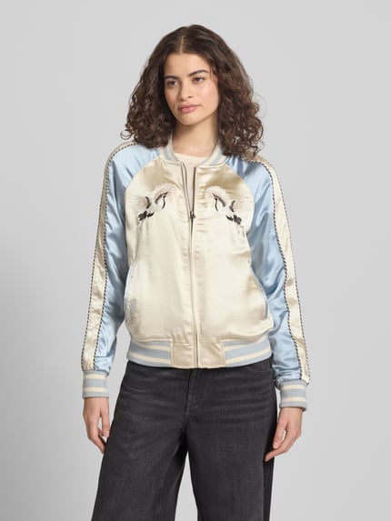 Frauenmodel trägt Bomber Jacke mit Motiv- und Logo-Stitching von Superdry aus Satin in hellem Blau und Beige mit Gerader-Schnitt, Stehkragen, Reißverschluss, Seitliche Eingrifftaschen und Elastische Abschlüsse. Nahaufnahme Oberkörper.