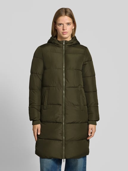 Frauenmodel trägt Puffermantel mit Kapuze von PIECES in Olivgrün, Longline-Form, Reißverschluss, seitliche Taschen, Ganzkörperansicht von vorne.