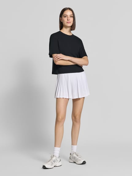 Frauenmodel trägt Relaxed Fit T-Shirt aus Baumwoll-Mix von Fabletics in Schwarz, kombiniert mit weißem Rock, Ganzkörperansicht von vorne.