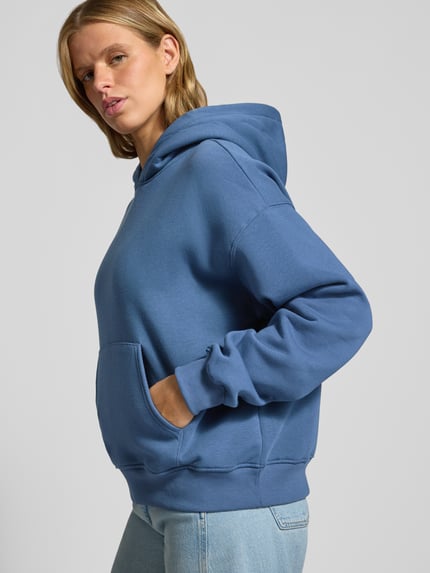 Frauenmodel trägt Hoodie mit Känguru-Tasche von Gina Tricot, Baumwoll-Mix, Relaxed Fit, überschnittene Schultern, Ganzkörperansicht von vorne.