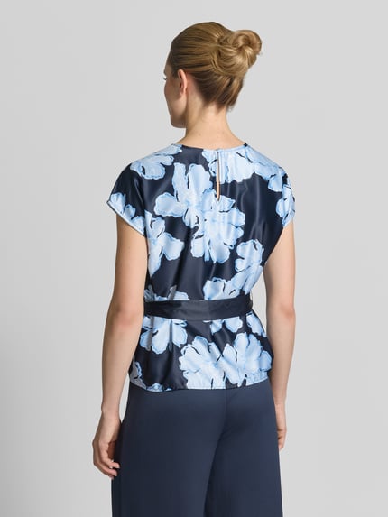 Vrouwenmodel draagt Blusenshirt aus Satin mit Bindegürtel van Christian Berg Cocktail, donkerblauw met grote lichtblauwe bloemen, getailleerd model, all-over look, strikceintuur, ronde hals, ruglengte maat 36: 58 cm, mouwlengte maat 36: 13 cm, satijn, dames, blouse met korte mouwen, geheellichaamsaanzicht achterkant.
