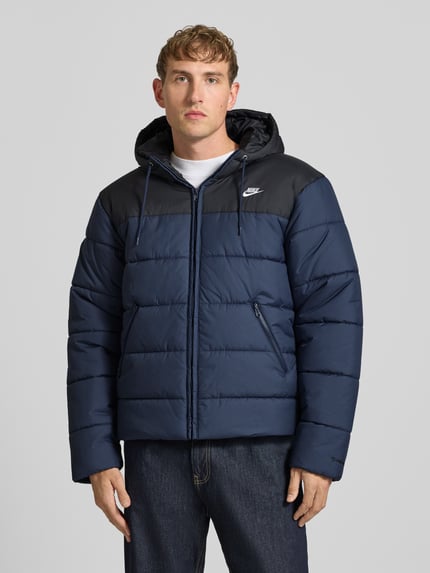 Männermodel trägt Steppjacke mit Kapuze von Nike in Two Tone Optik mit Struktur, Reißverschlusstaschen und Logo-Print, Ganzkörperansicht von vorne.