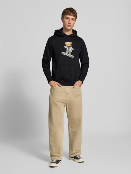 Männermodel trägt schwarzen Relaxed Fit Hoodie aus Baumwoll-Mix mit Motiv-Print von CHILL PILL, Känguru-Tasche, elastische Abschlüsse, Ganzkörperansicht von vorne.