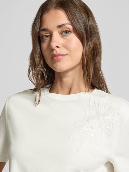 Frauenmodel trägt Regular Fit T-Shirt aus reiner Baumwolle mit Blumen-Applikation von Christian Berg Woman, cremeweiß, Nahaufnahme Oberkörper. Rückenlänge bei Größe S 54 cm, Pailletten-Details, Rundhalsausschnitt, überschnittene Schultern.
