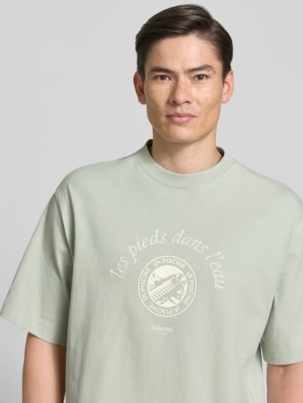 Männermodel trägt Relaxed Fit T-Shirt aus reiner Baumwolle Modell 'BOXYNORMAN GRAPHIC' von SELECTED HOMME in hellem Grün, Motiv-Print, Nahaufnahme Oberkörper.