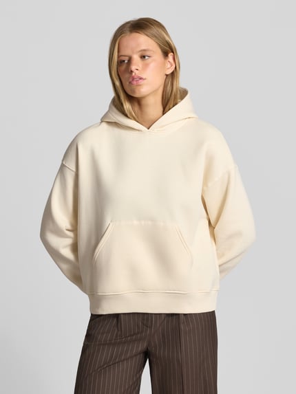 Modelka nosi Relaxed Fit Hoodie aus Baumwoll-Mix Modell 'Iconic' von Gina Tricot, kolor ecru, kieszeń kangurka, Mieszanka bawełny, wykończenia elastyczne, całkowity widok z przodu.