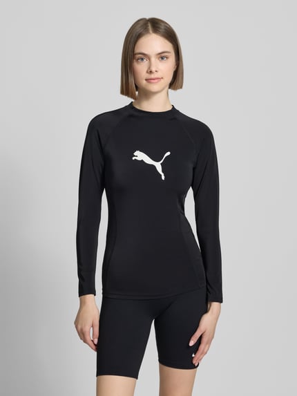 Vrouwenmodel draagt Badeshirt im langärmeligen Design Modell 'RASH GUARD' van PUMA, zwart, recht model, materiaalmix met stretch, ronde hals, logoprint, uv-bescherming 50+, chloorbestendig, ruglengte maat S 56 cm, voorkant bovenlichaam close-up.