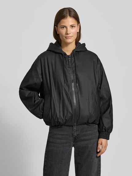 Kobiece model prezentuje czarną, luźną kurtkę z kapturem Jacke mit Kapuze Modell 'LOHJA' od RAINS, matowa faktura, zamek, kieszenie, elastyczne mankiety, widok całej sylwetki z przodu.