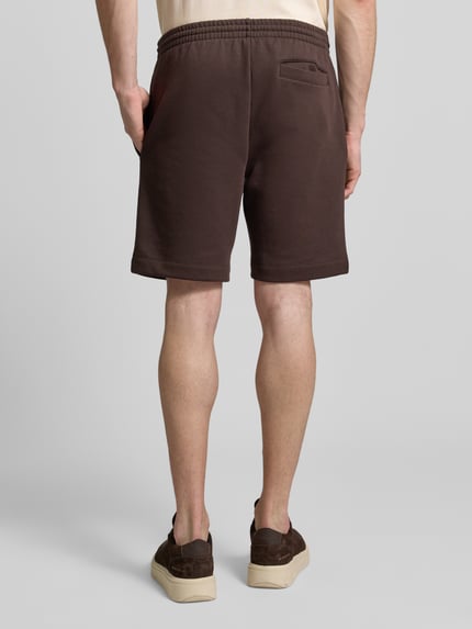 Männermodel trägt Regular Fit Sweatshorts aus Baumwoll-Mix von Lacoste, unifarben braun, elastischer Bund, seitliche und Gesäßtaschen, Ganzkörperansicht Rückansicht.