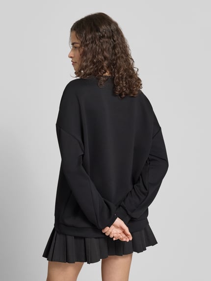 Młodzieżowy model nosi Relaxed Fit Pullover mit Herz-Stickerei Model 'TALIA' od Pieces, efekt: jednolity kolor, krój relaxed fit, wyhaftowane serce, okrągły dekolt, długi rękaw, czarny, tkanina gładka, całkowity widok z tyłu.