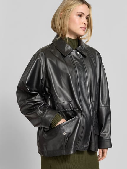 Frauenmodel trägt unifarbene, schwarze Funktionsjacke Modell 'GROOVE' von IRO in Lederoptik mit metallischen Akzenten, Oversized, Longline, Kapuze, Taschen, glänzende strukturierte Oberfläche, Nahaufnahme Oberkörper.