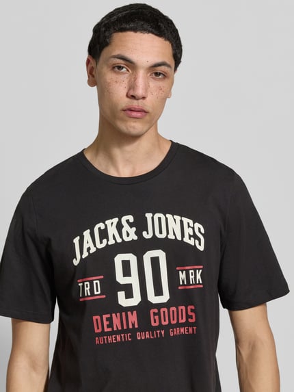 Mężczyzna nosi T-Shirt mit Label-Print im 3er-Pack Modell 'ETHAN' von Jack & Jones z czarnej bawełny, prosty krój, okrągły dekolt, nadruk z przodu, kontekst Herren T-Shirt z długością tyłu 67 cm w rozmiarze M, zbliżenie na górną część ciała.