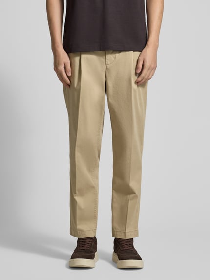 Männermodel trägt Regular Fit Chino aus Baumwoll-Mix Modell 'TAAMIR PLEATED' von Armedangels in Beige mit Logo-Stitching, Ganzkörperansicht von vorne.