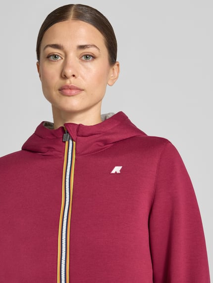 Frauenmodel trägt Sweatjacke mit Futter aus Kunstfell und Zweiwege-Reißverschluss von K-WAY, bordeauxrot, Logo, Kapuze, Nahaufnahme Oberkörper. Material-Mix mit Stretch-Anteil, Regular Fit, sichtbarer Reißverschluss, dezente Textur.