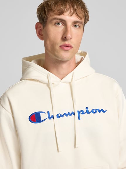 Männermodel trägt weißen Hoodie mit Label-Stitching von Champion aus Baumwoll-Mix, lockerer Schnitt, Nahaufnahme Oberkörper.