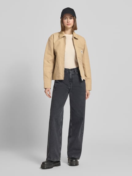 Frauenmodel trägt Jacke mit Label-Detail von Carhartt Work In Progress in Beige aus Reine Baumwolle, Relaxed-Fit, Umlegekragen, Reißverschluss, Eingrifftaschen, kombiniert zu weiter Jeans und Beanie, Rückenlänge bei Größe S: 58 cm, Schulterbreite: 47 cm, Brustweite: 54 cm, Ärmellänge: 60 cm, Ganzkörperansicht von vorne.