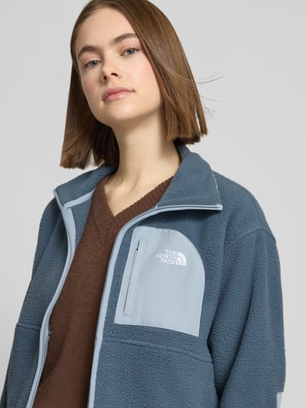 Frauenmodel trägt Fleecejacke mit Stehkragen und Label-Stitching von The North Face in Blau mit Reißverschluss und Brusttasche, Nahaufnahme Oberkörper.